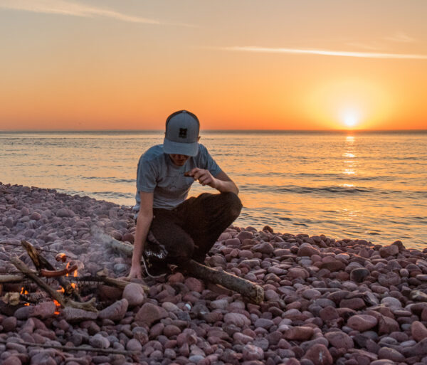 Young man tending a fire on rocky beach with sun setting